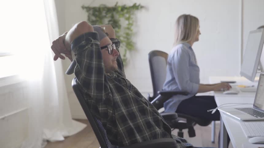 A man leans back in his chair while a woman works at her computer while they both sit in front of a long white desk with a large window behind them