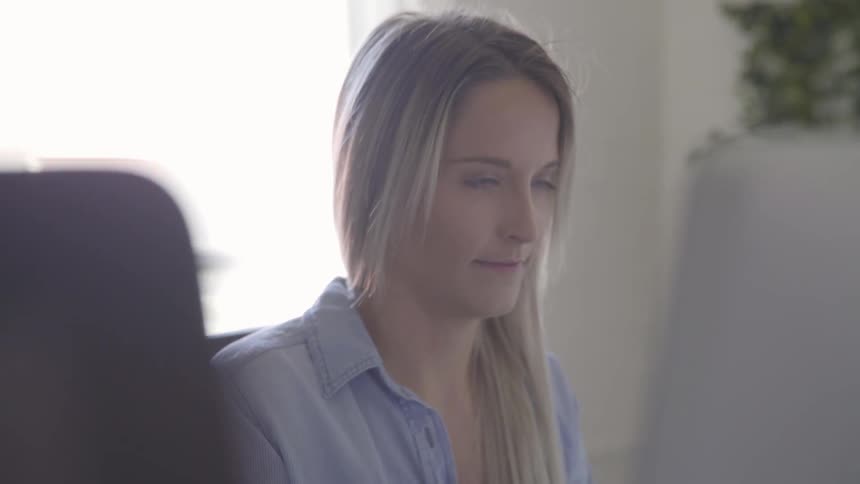 A blonde female sits at a desk and works on a desktop computer in a bright office
