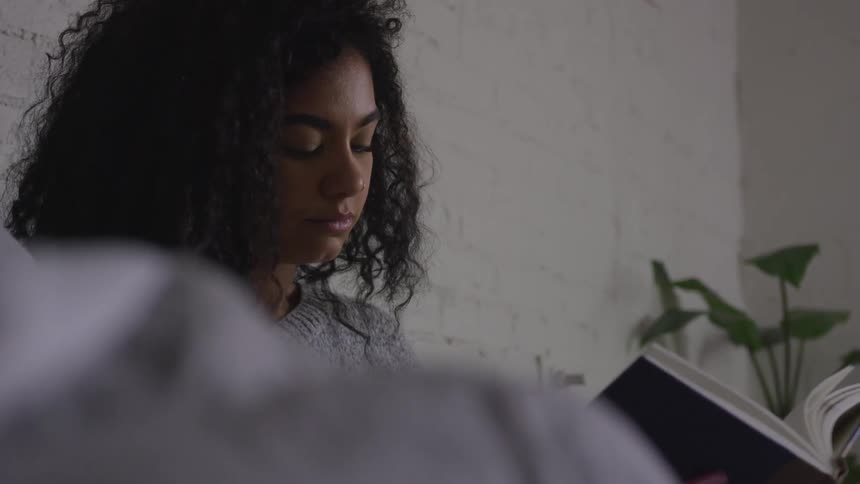 A young adult female sits reading a book in front of a white brick wall with a plant in the corner