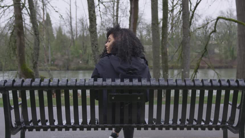 A young female sits on a park bench with trees and a river in front of her