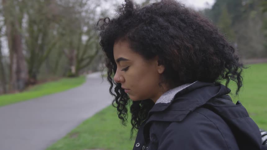 A young black woman wearing a black raincoat stands outside in an area with grass and trees next to a path as she looks deep in thought