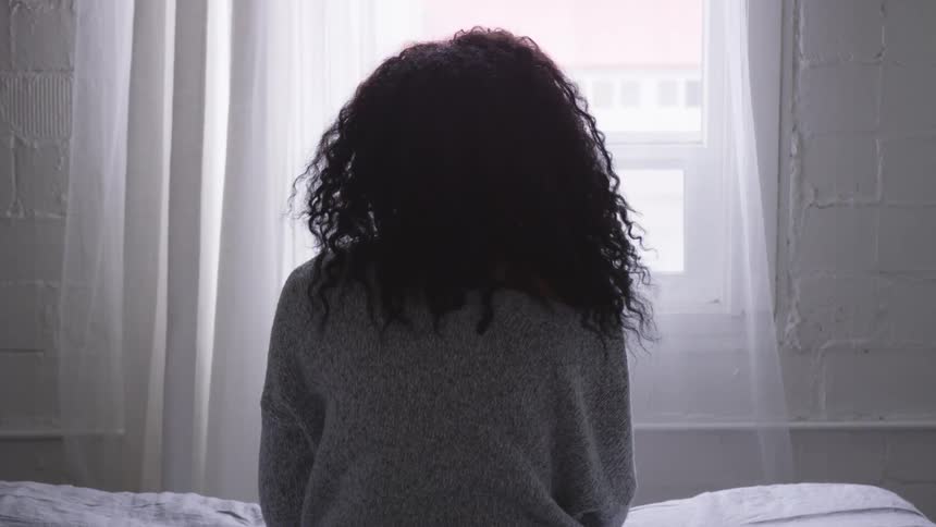 A woman sits on a bed and stares out a window