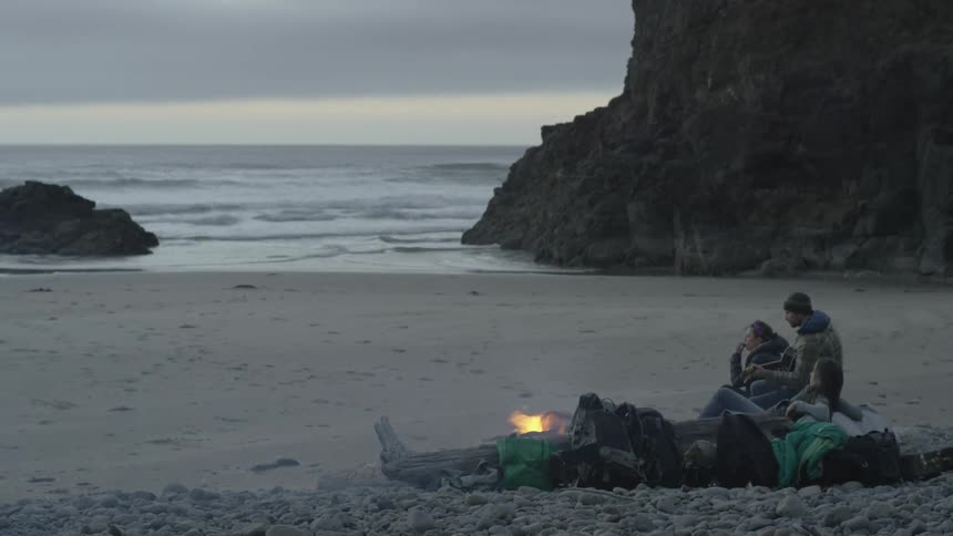 A man and woman converse while seated next to a campfire on the beach