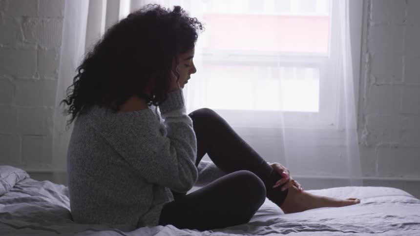 A young woman sits on a bed and puts her hand to her chin