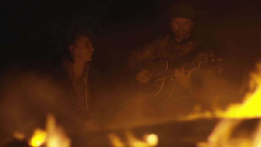 A man plays guitar and sings for friends around a campfire on the coast enjoying life