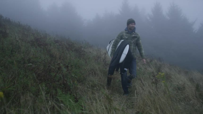 A man carrying a surfboard through tall grass on a hillside on a foggy day with strong wind blowing the grass