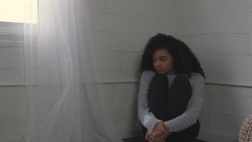 A woman sits in the corner of a white room with her arms wrapped around her legs