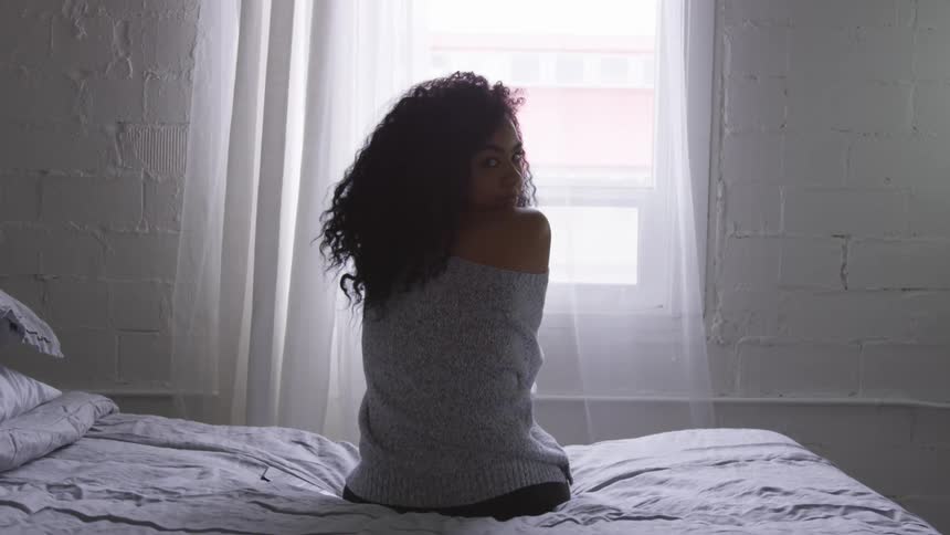 A black woman sits on a bed and turns her head around to stare
