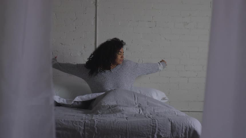A young adult female wearing a gray sweater stretches her arms wide while sitting in a bed in front of a white brick wall