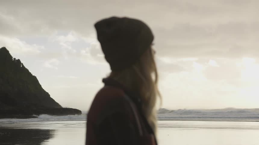 A woman wearing a beanie stands on the beach and stares out towards the ocean