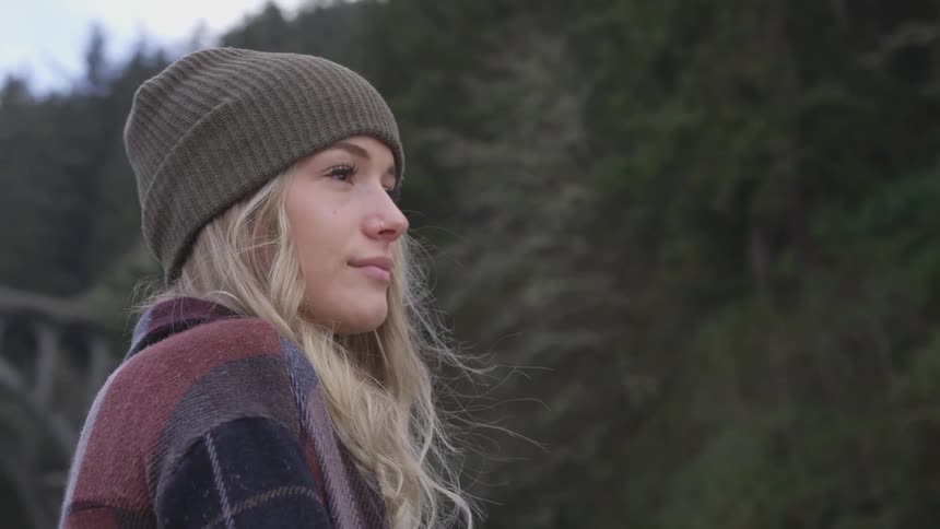 A white woman outdoors wearing a knit beanie slightly smiles as she looks off into the distance