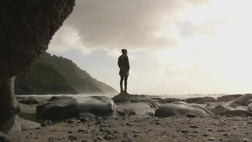 A person stands on a rock at a beach and stares out into the ocean