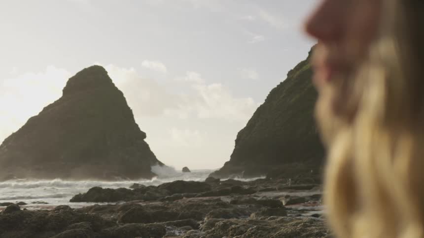 A lovely young woman looks off in the distance as she stands on a rocky coastline upon which waves are violent crashing