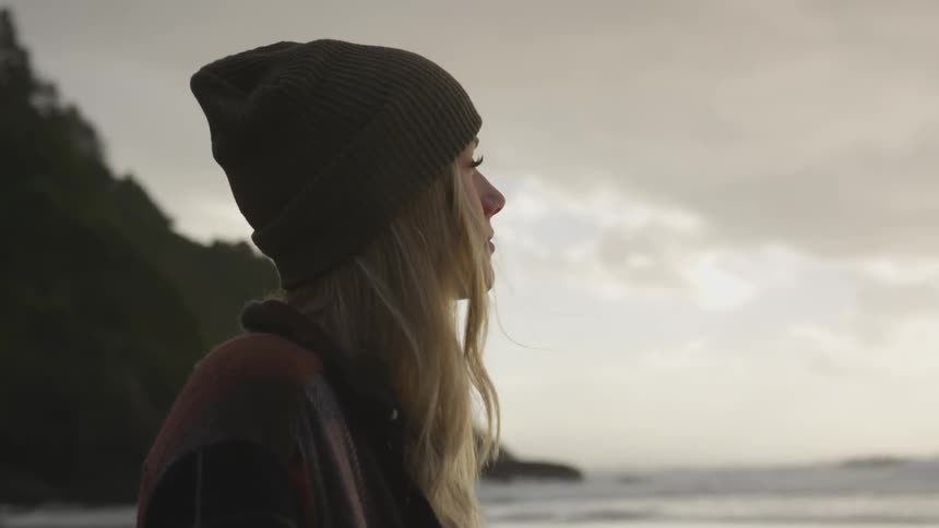 A young woman with long hair and wearing a knit hat stands on a beach at the foot of a forested mountain and she looks out over the ocean in front of her