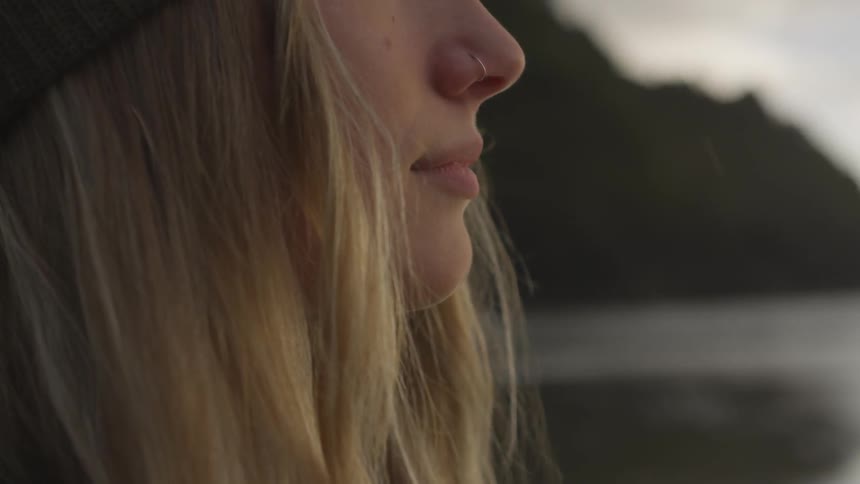 Blonde woman wearing wooly hat and nose ring looks ahead outdoors at dusk