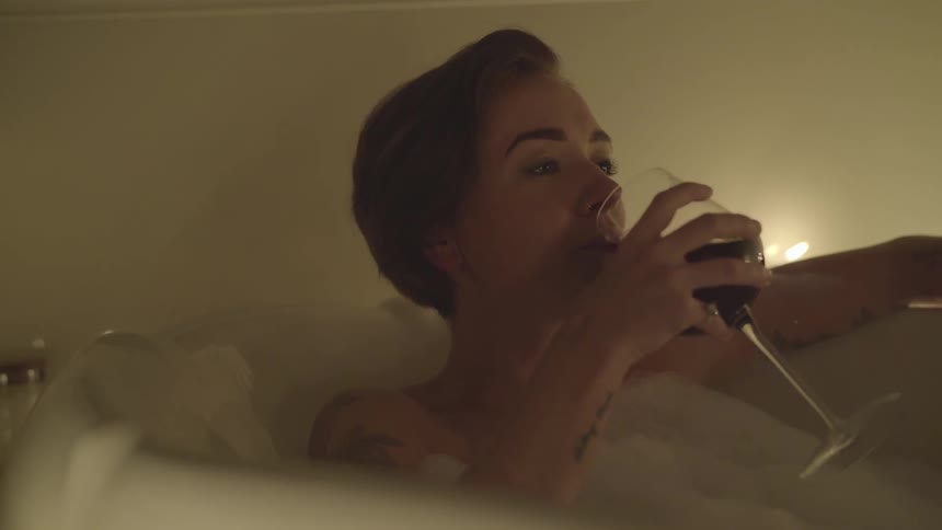 A woman has a bath in candle light and sips red wine