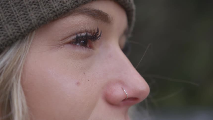 A blonde woman with a nose ring and wearing a winter hat stands outside and blinks as she looks