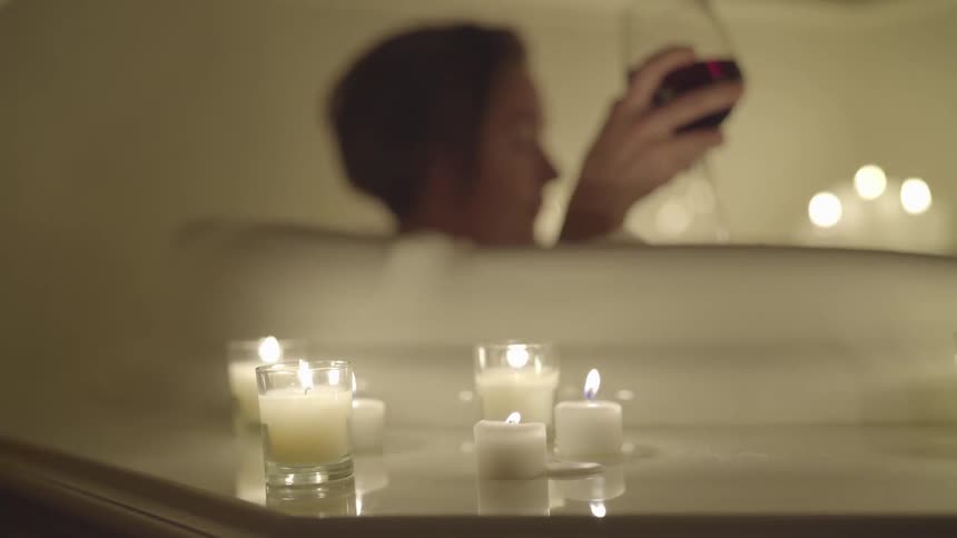 A woman drinks a glass of wine in a bathtub surrounded by candles