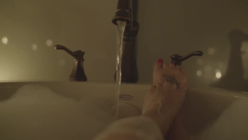 Woman taking a bubble bath rubs her feet together with candles in the background