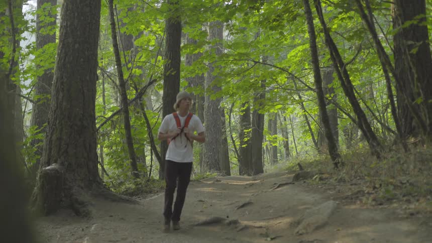 A white teenager wearing a hat and a backpack hikes along a forest path