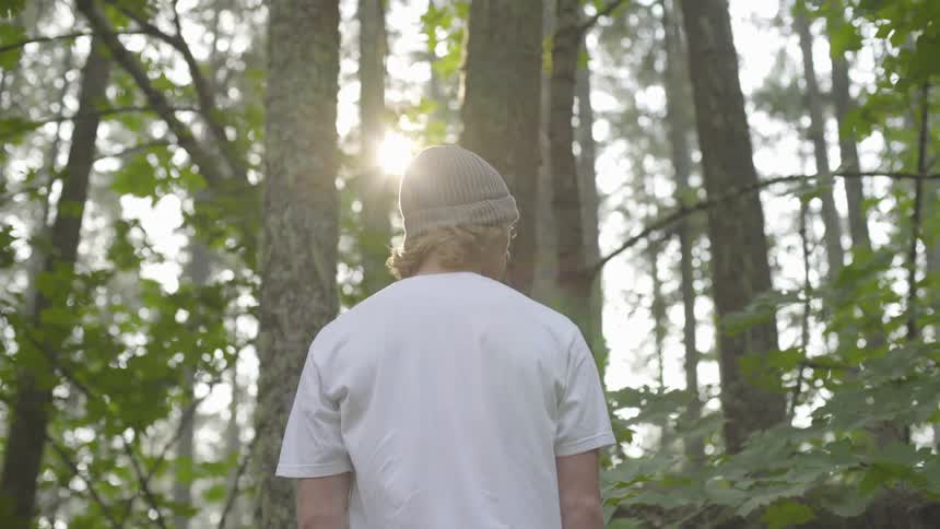 Man wearing white t shirt and wooly hat stands in the forest while the sun shines on his head