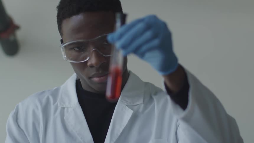 A black man in a lab coat swishes a red fluid inside a test tube