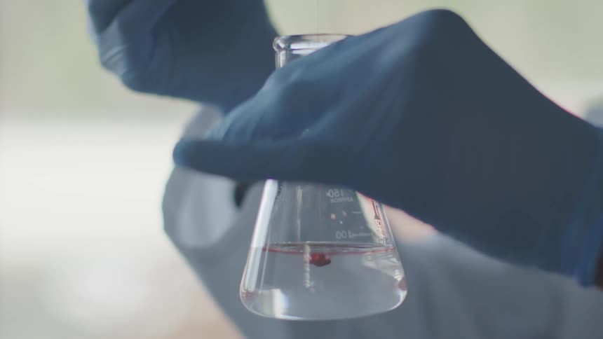 A lab worker drops blobs of a red liquid into a beaker partially filled with a cloudy fluid
