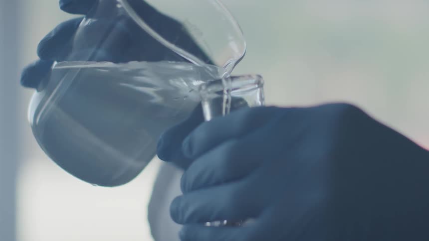 A person wearing gloves pours clear liquid from a large beaker into a smaller beaker