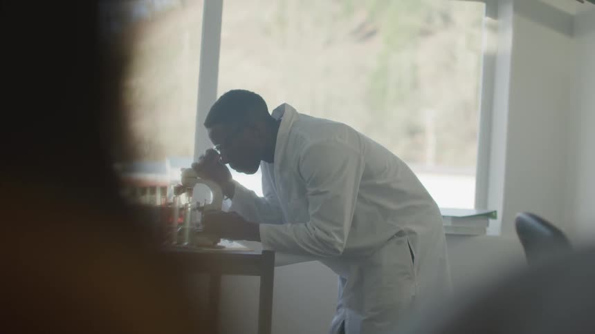 A man in a lab coat looks into a microscope that's sitting on a table