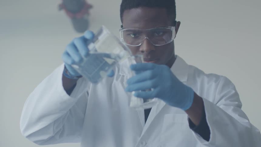 A young black man in a lab coat pours liquid into a beaker
