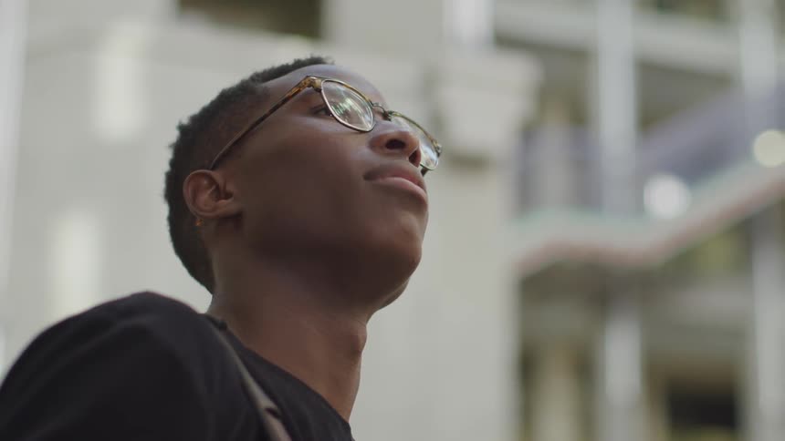 A black man wearing eyeglasses looks upward towards the sky while standing in front of a large white building with many windows