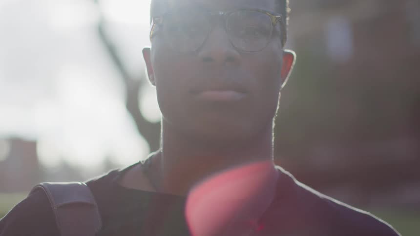 A young black man in glasses is standing outside as the sun shines brightly behind his head