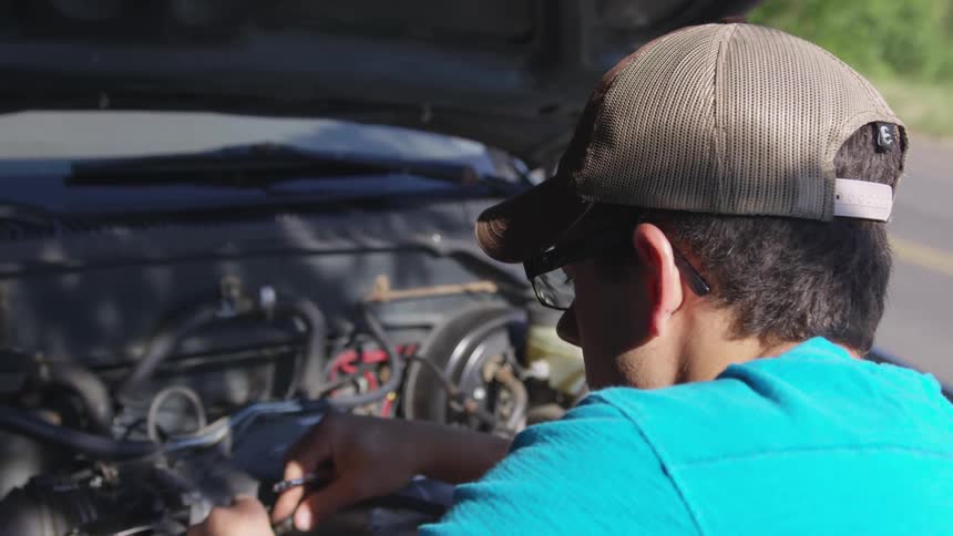 A bespectacled man in a cap works on an engine on a sunny day