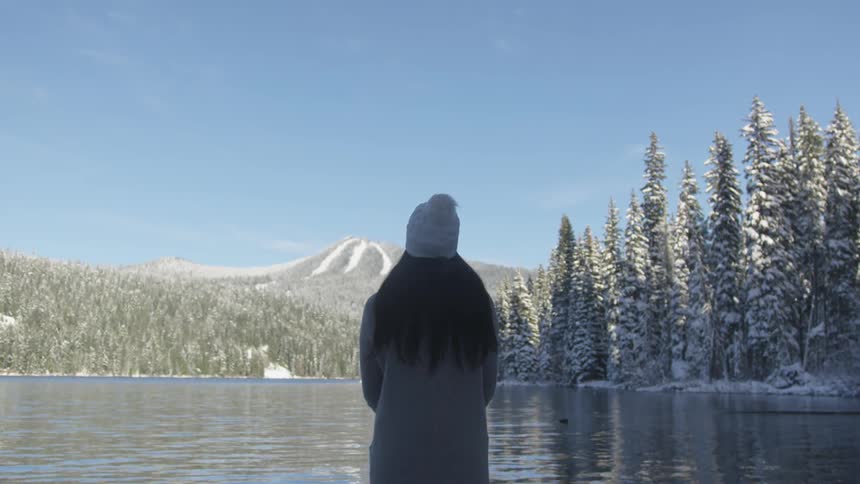Long black hair woman wearing wooly hat stands in front of snowy forest lake in the daytime
