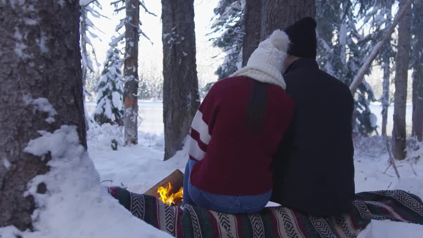 A man and a woman are sitting close to an outdoor campfire in the snowy woods