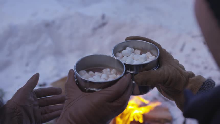 Two campers toast each other with cups of cocoa with floating marshmallows in front of a flaming fire in the snow