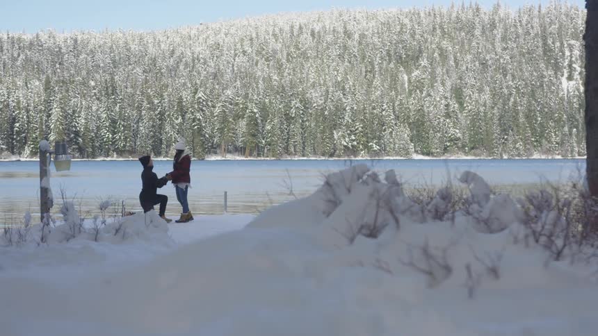 A person kneels in front of another person in front of water in the winter