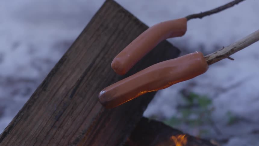 Two hot dogs are being grilled on a campfire with a stick in a snowy area