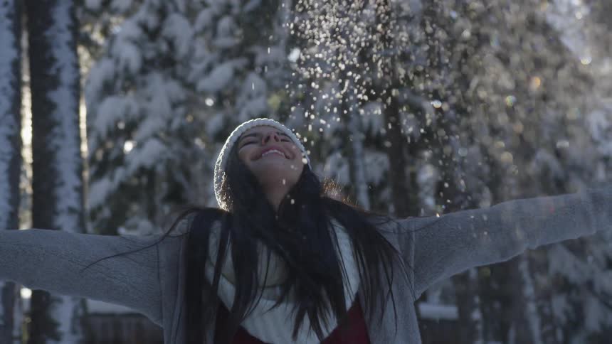 Woman wearing white wooly hat and scarf throws snow up into the air during at sunset