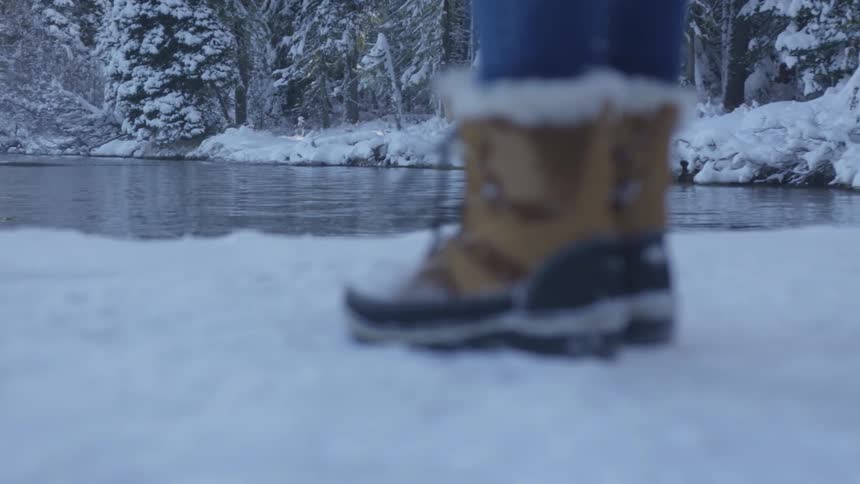 A person wearing fur topped boots stands on the shore of an icy lake