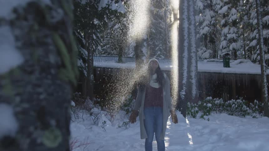 A woman in a knit hat and coat stands in front of a secluded cabin in a forest that is coated in snow and she takes a handful of snow and throws it in the air and smiles as more snow continues to fall from the sky