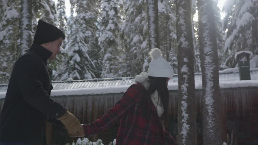 A young woman pulling a young man along by the hand in front of a cabin in a snowy forest