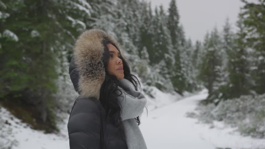 A woman wears a heavy coat and looks at the snow topped forest trees