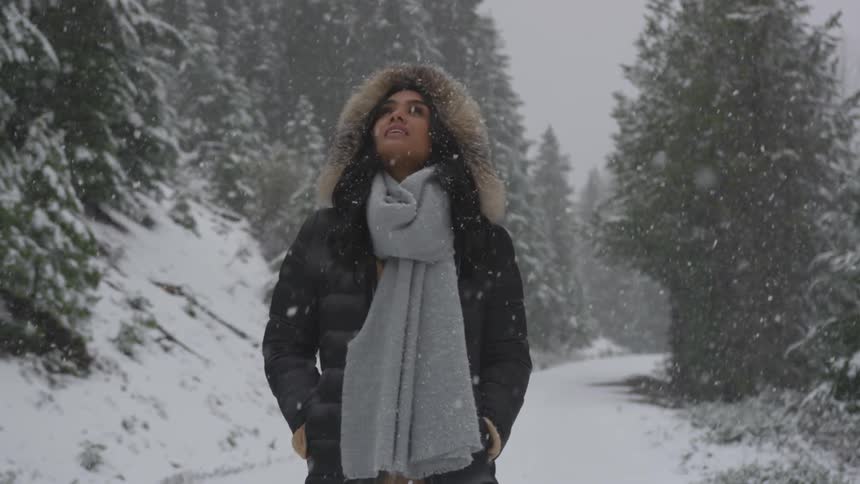 A woman in a heavy coat walks outside in a forest while it snows