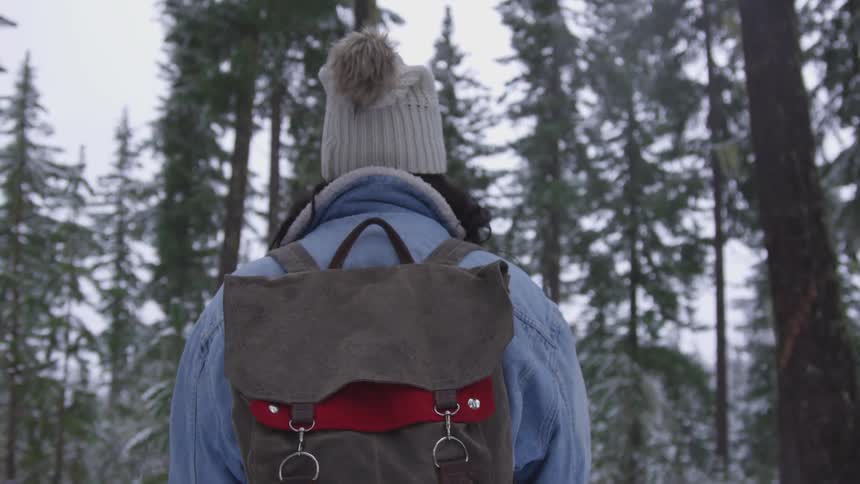 A person in a bobble hat and denim jacket wears a backpack while walking through a forest in the snow