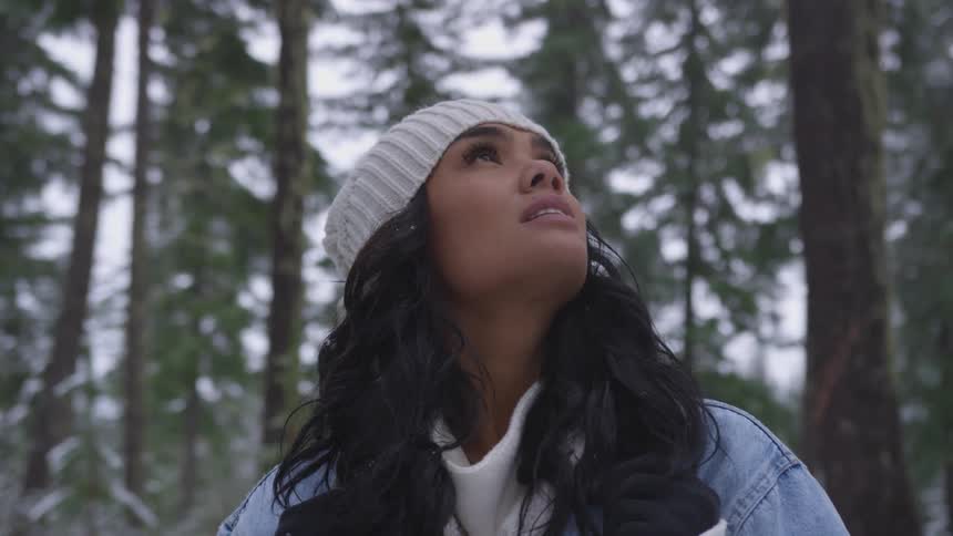 A woman wearing a white knit hat looks up through the pine trees as a light snow starts falling down around her