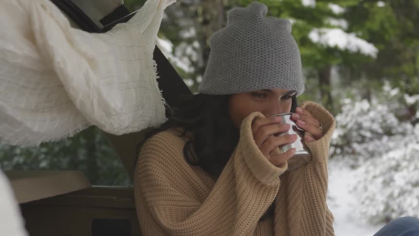A woman sits outside on the back of her car drinking