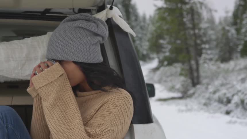 A woman sits on the back of a car and sips a warm drink on a cold day
