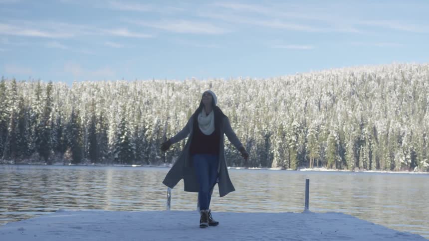 A woman twirls around in the snow next to a lake