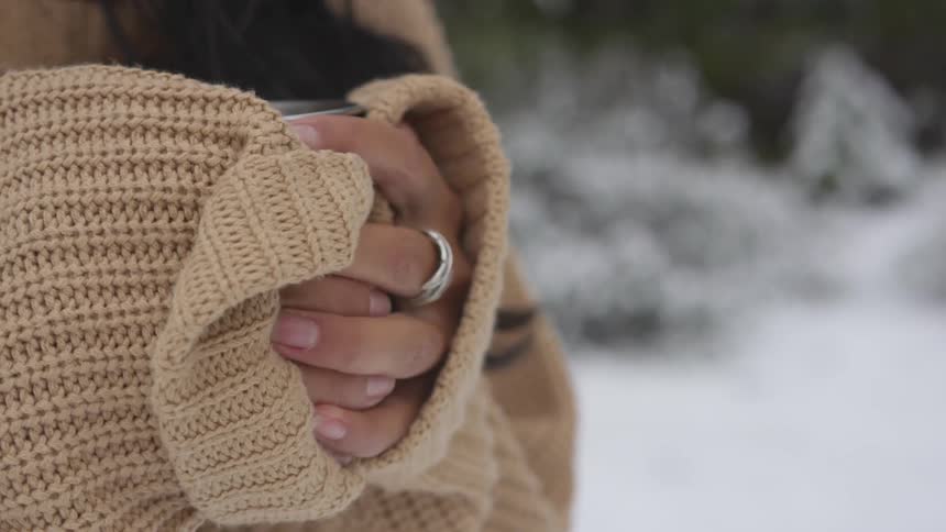 Woman stands outside in snow clasping hands around cup